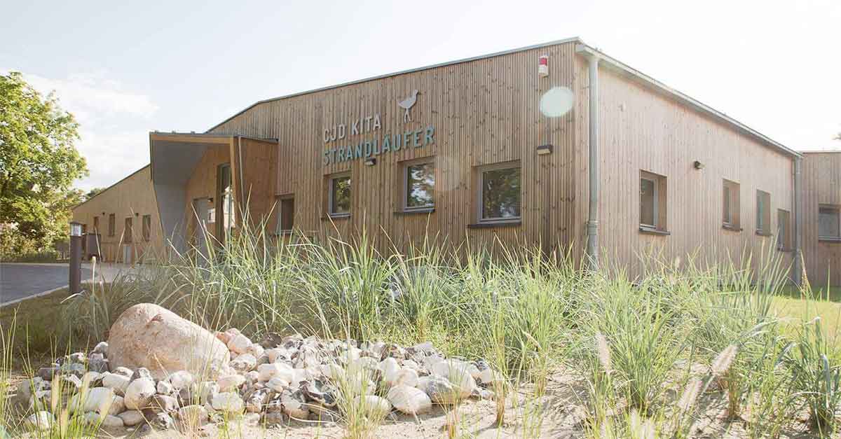 Holzgebäude mit Satteldach, versetzt angeordneten Fenstern und asymmetrische Überdachung am Eingang. Schriftzug an der Fassade: CJD Kita Strandläufer.