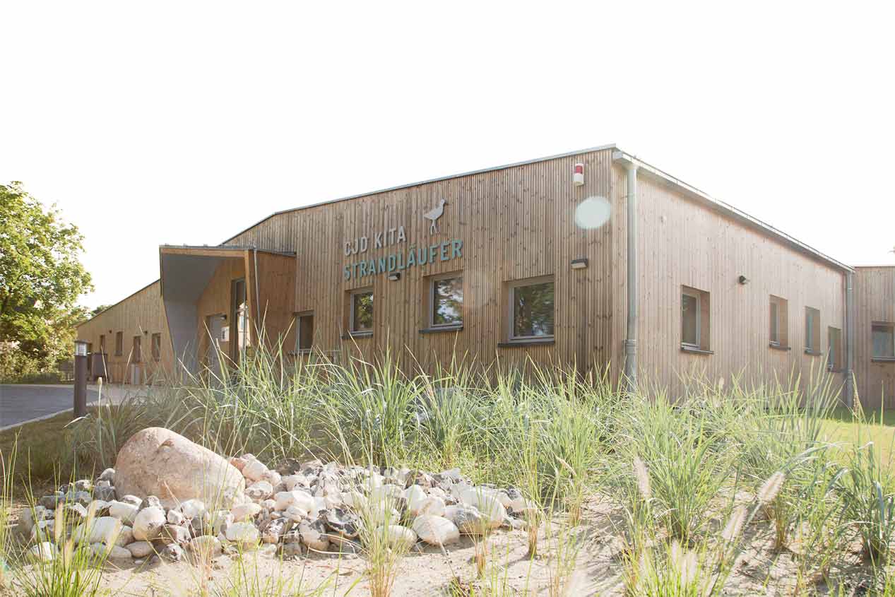Holzgebäude mit Satteldach, versetzt angeordneten Fenstern und asymmetrische Überdachung am Eingang. Schriftzug an der Fassade: CJD Kita Strandläufer.