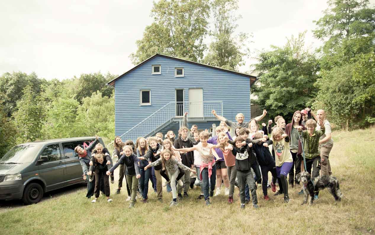 Foto Schulgruppe Montessori. Ausgelassene Stimmung. Im Hintergrund blaues Holzhaus und Umgebung.