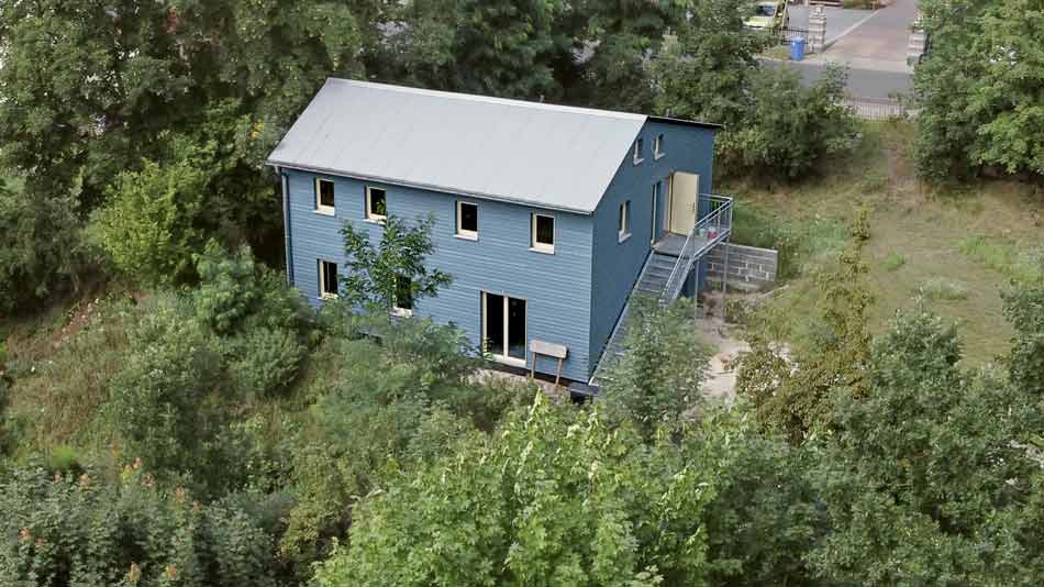 Foto in Vogelperspektive des "blauen Hauses". Blick auf gesamtes Holzgebäude mit außen liegender Stahltreppe und umliegender Vegetation.