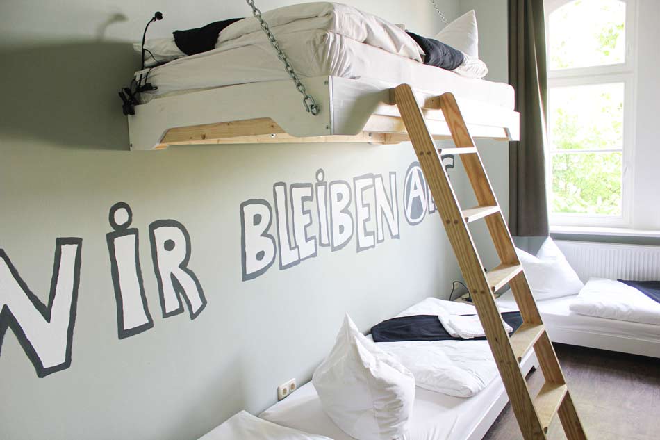 Ein Zimmer des Postels in Wolgast. Zu sehen sind mehrere Betten in weiß gehalten, und eine grün-mint farbene Wand auf der in Großbuchstaben geschrieben steht: Wir bleiben alle.