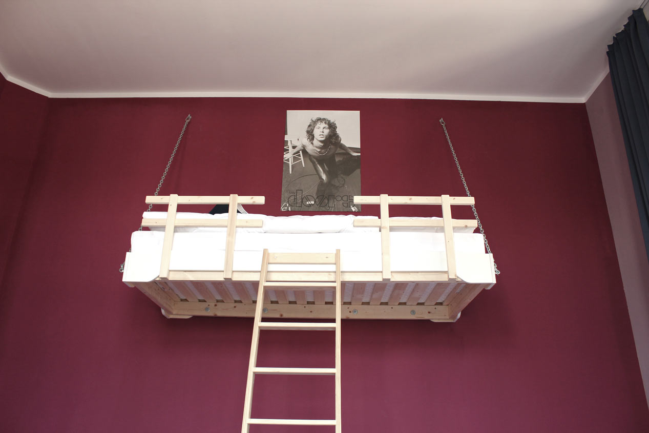Ein Zimmer des Postels in Wolgast. Ein Hochbett mit Holzleiter an einer brombeer-roten Wand, darüber ein Poster des Sängers von der Band The Doors.