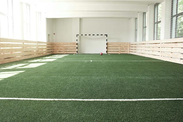 Indoor Fußballfeld im Postel Wolgast, mit Kunstrasen, einem Tor, einem Ball, vergitterten Fenstern und Holzlatten als Feldbegrenzung.
