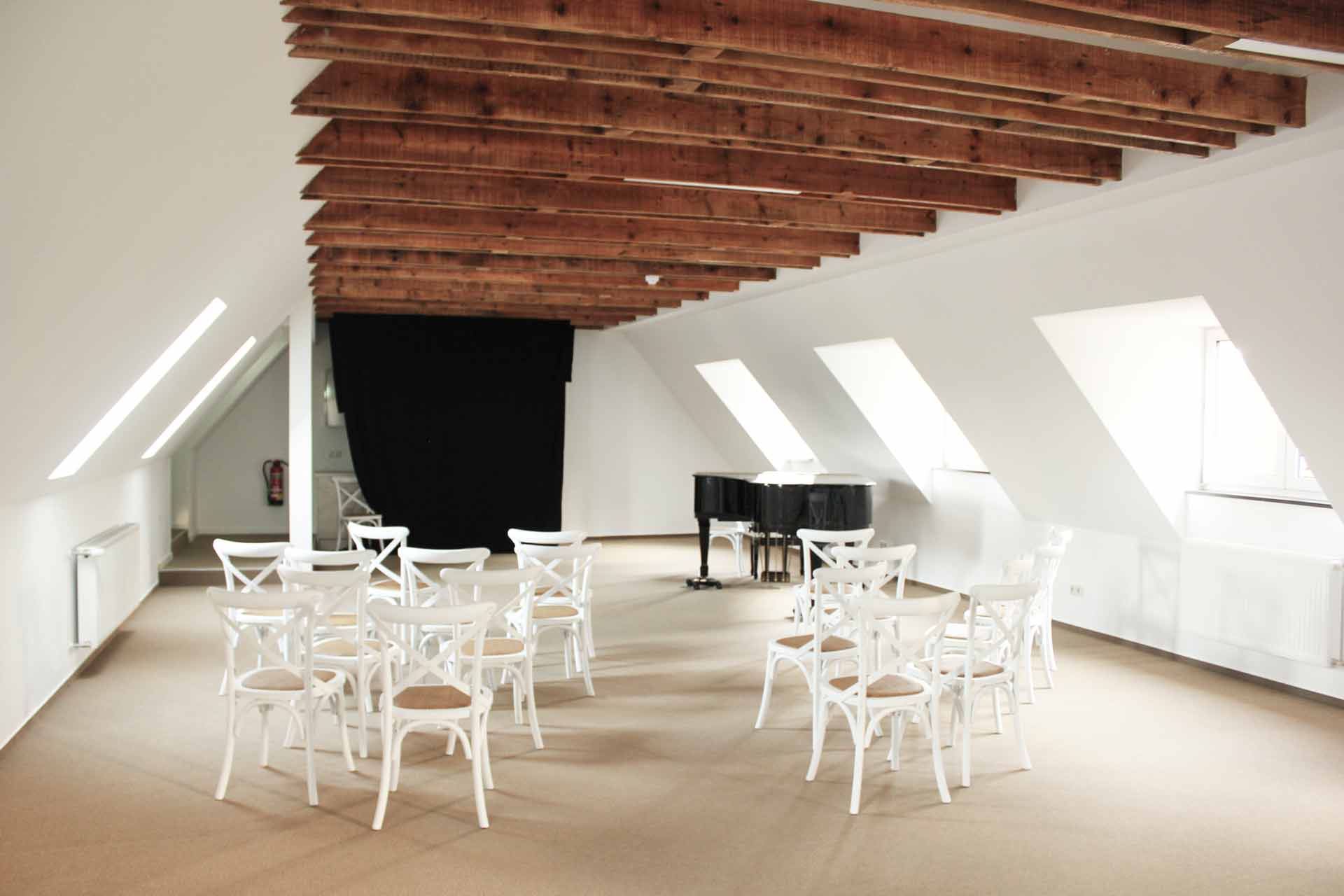 Großzügiger Raum für musikalische Veranstaltungen im Postel Wolgast. Ein Klavierflügel, mehrere weiße Stühle und viele sichtbare Holzbalken an der Decke machen den Charme des Raums aus.