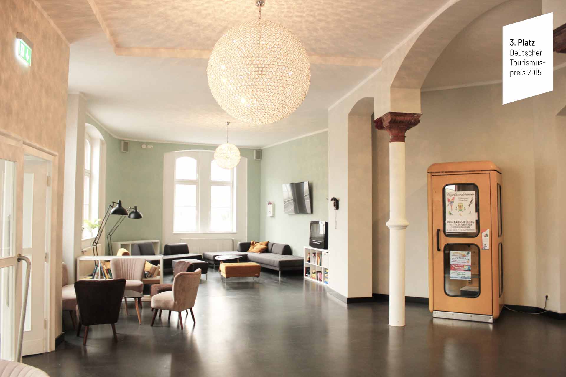 Foyer im Postel Wolgast bei Tageslicht, mit Tischen und Stühlen, einer angeschalteten Lampe das Muster an die Decke wirft und einer Telefonzelle. Darauf ist ein weißes Feld mit der Aufschrift "3. Platz Deutscher Tourismuspreis 2015"