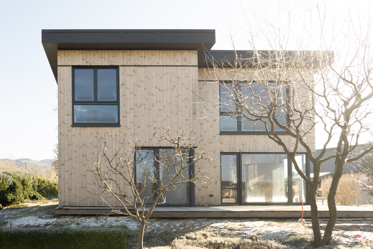Foto RING Fassadenansicht. Man sieht die helle Holzfassade des Gebäudes sowie die dunklen Fenster. Das Dach ist ebenfalls aus dunklem Metall und ist als Flachdach ausgeführt.