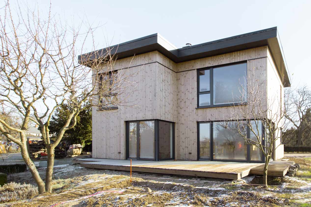 Bild von Holzhaus mit heller Holzfassade. Das Haus ist zwei geschossig und verfügt über ein Flachdach. Die Fenster sind groß und haben einen dunklen Rahmen. Im Vordergrund ist eine Terrasse angelegt.