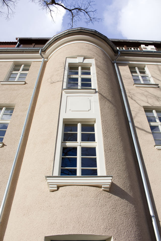 INFA-Detail Die Fassade des Mannschaftsgebäudes des INFA-Geländes ist zu sehen. Das Gebäude ist beige verputzt und hat weiße mehrfachgeteilte Fenster.