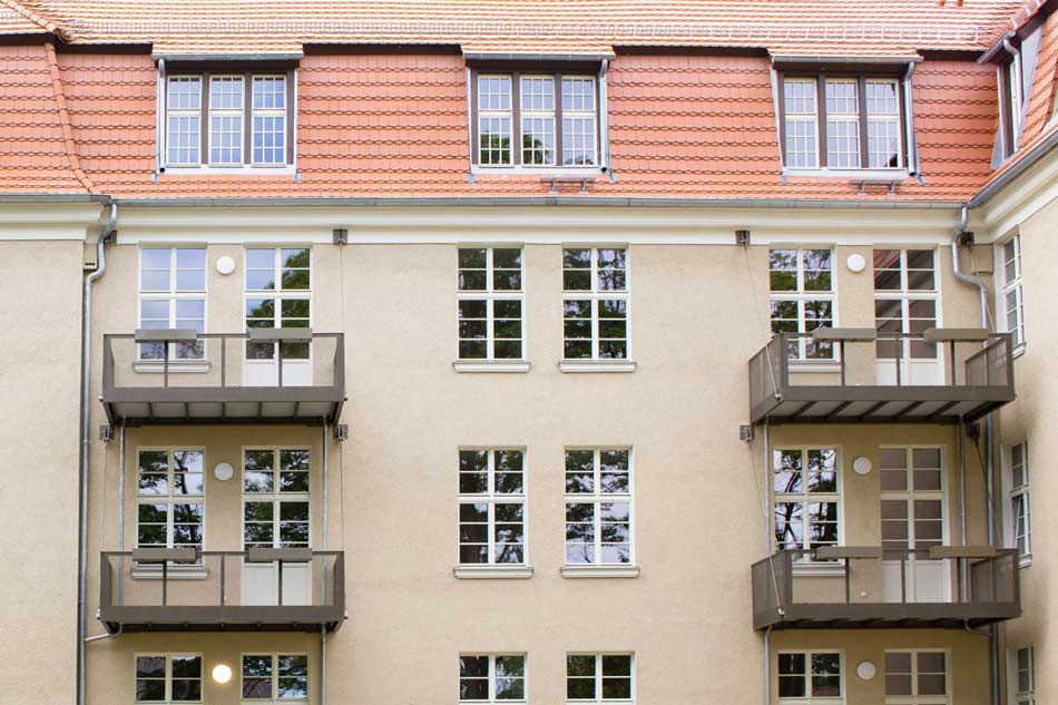Nahaufnahme auf Balkone an der Ostfassade des Wohngebäudes (ehemaliges Mannschaftsgebäude) im INFA Quartier. Das Gebäude ist beige verputzt und hat weiße mehrfachgeteilte Fenster.