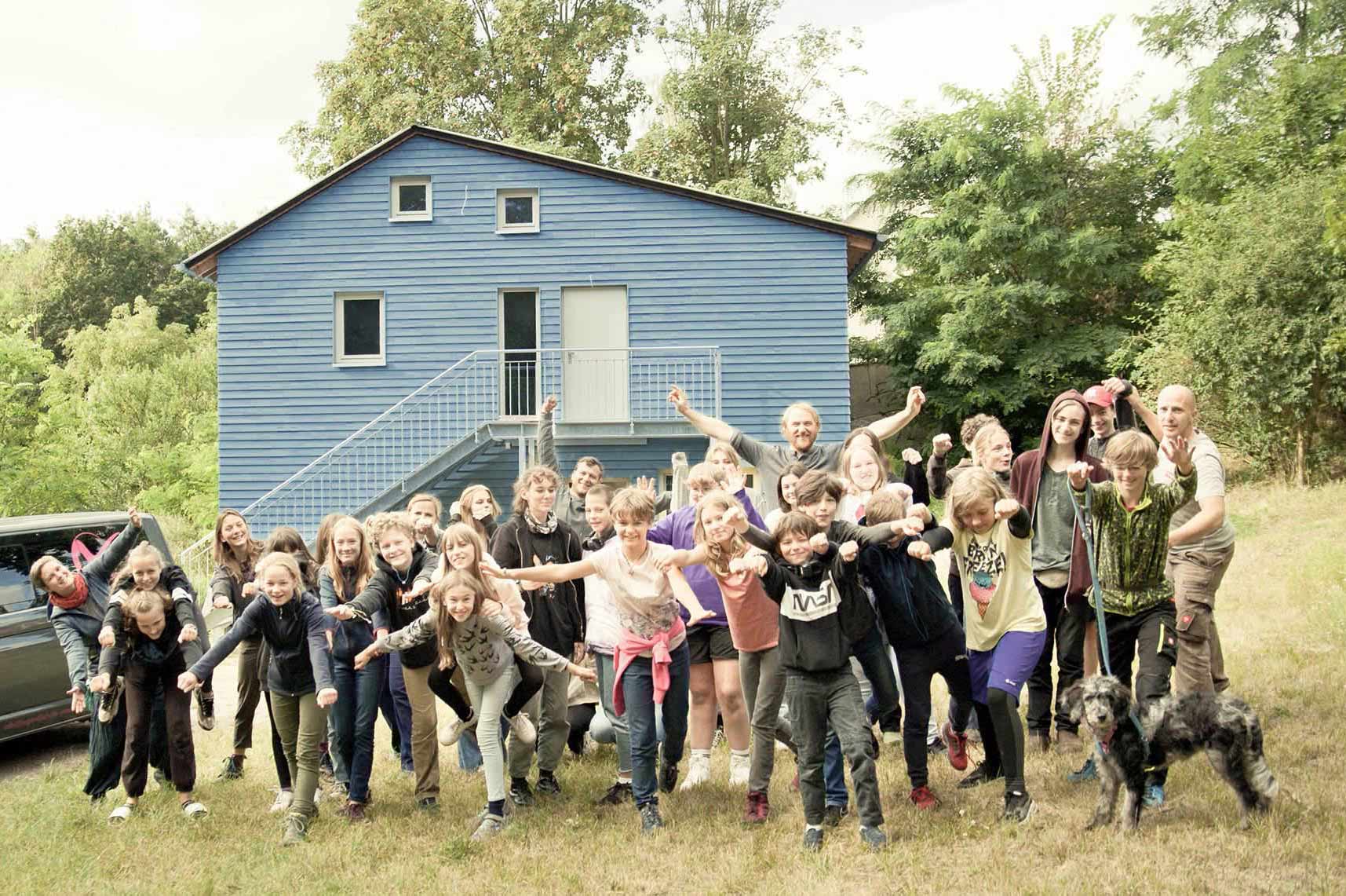 HENN_cubus-strausberg-blaueshaus-1700 Foto Schulgruppe Montessori. Ausgelassene Stimmung. Im Hintergrund blaues Holzhaus und Umgebung.