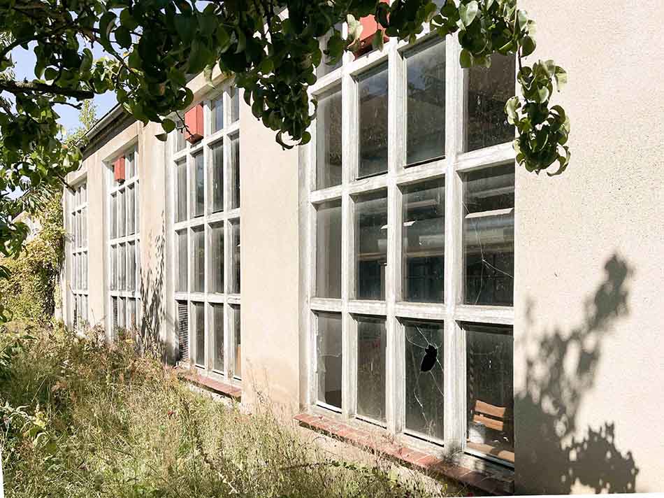 Jugendclub-Außenansicht_Fensterdetail Außenansicht des geplanten Jugendclubs, mit Fensterdetail, die Glasscheiben sind teilweise gesprungen und haben Einschläge, hell verputzte Wand