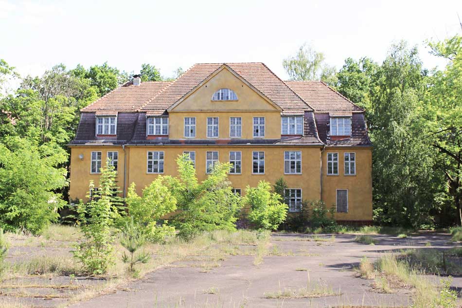 das ehemalige Stabshaus auf dem Gelände der ehemaligen Infanterieschule Wünsdorf in ockerfarbenem Putz, davor Vegetation auf einem maroden Vorplatz.