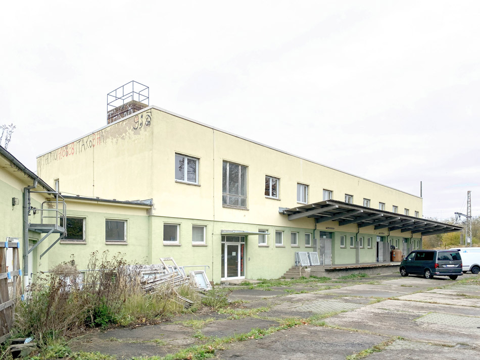 BAHN5-2_cubus-plan Die Vorderseite des Gebäudes vor dem Umbau mit gelb-grüner Farbe und Graffiti im oberen Fassadenbereich und noch nicht barrierefrei. Unkraut wächst in den Plattenfugen vor dem Gebäude und Fensterrahmen stehen angelehnt.