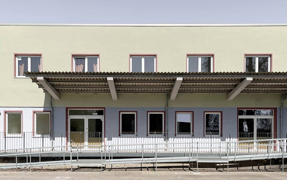 BAHN5-4_cubus-plan Angeschnittene frontale Ansicht des EFJ-Wohngebäudes mit zweifarbiger Zonierung, unten sanftes Blaugrau oben helles Grün. Rollstuhlzugang, barrierefreier Eingang des Umbaus.