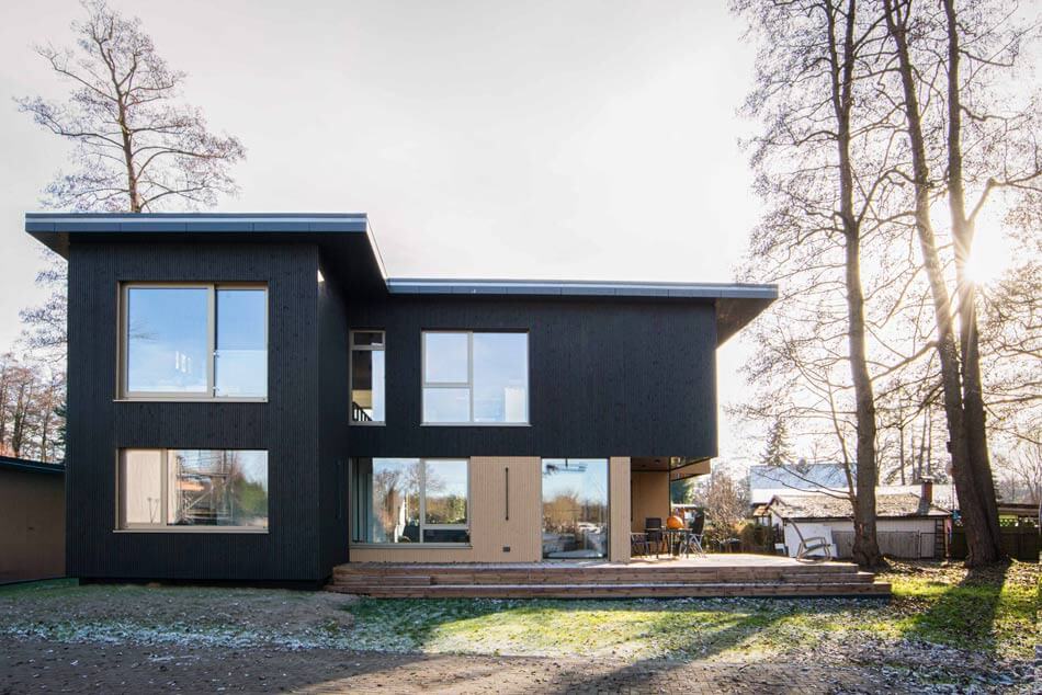 Das Holzhaus mit dunklem und hellem Holz und einer Terrasse mit Möbeln. Rechts unter hinter dem Haus stehen Bäume, ringsherum ist Wiese und die Sonne scheint auf den leicht angeschneiten Boden.