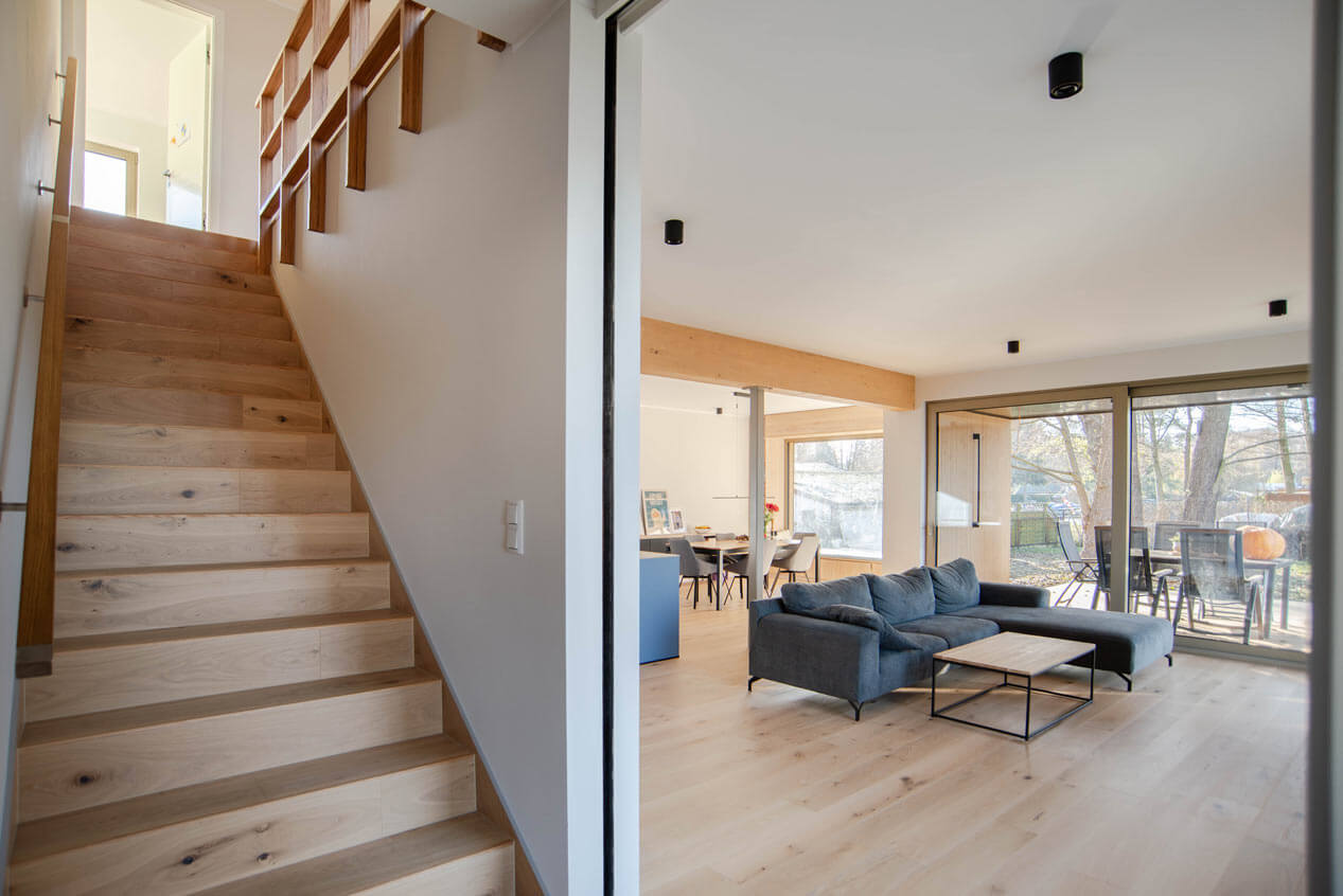 Blick in den Wohn- und Ess- und Küchenbereich mit Tisch, Stühlen und Sitznischenfenster im Hintergrund. Mittig blickt man in den Wohnbereich, mit einer grauen Couch und Wohnzimmertisch. Der Wohnzimmerbereich wird durch einen hellen Balken vom Ess- und Küchenbereich optisch getrennt. Große Schiebetürenfenster lassen auf die möblierte Terrasse blicken. Links ist der Treppenaufgang in das erste Obergeschoss zu sehen.