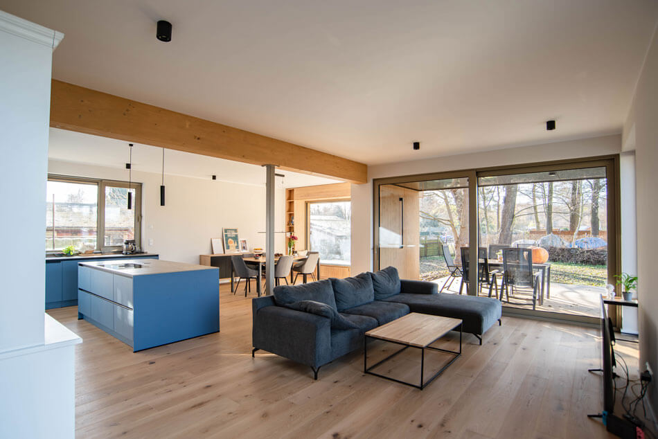 Blick in den Wohn- und Ess- und Küchenbereich mit Tisch und Stühlen im Hintergrund, links die Küche mit blauen Möbeln und mittig einer Kochinsel. Rechts davon blickt man in den Wohnbereich, mit einer grauen Couch und Wohnzimmertisch. Der Wohnzimmerbereich wird durch einen hellen Balken vom Ess- und Küchenbereich optisch getrennt. Große Schiebetürenfenster lassen auf die möblierte Terrasse blicken.