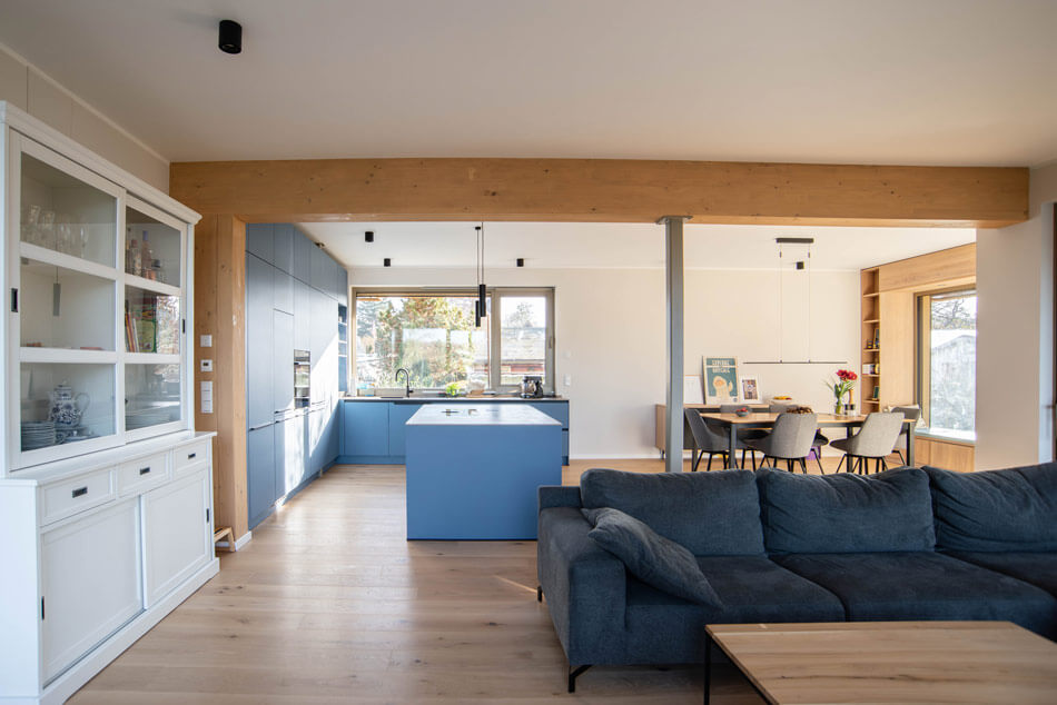 Blick in den Wohn- und Ess- und Küchenbereich mit Vitrine links, Tisch und Stühlen im Hintergrund, zentral die Küche mit blauen Möbeln und mittig einer Kochinsel. Rechts davon blickt man in den Wohnbereich, mit einer grauen Couch und Wohnzimmertisch. Der Wohnzimmerbereich wird durch einen hellen Balken vom Ess- und Küchenbereich optisch getrennt.
