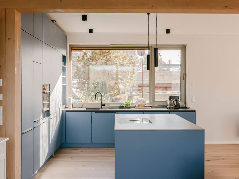 Blick in den Küchenbereich mit blauen Möbeln und mittig einer Kochinsel. Durch das große Fenster kann man in den Garten dahinter blicken.