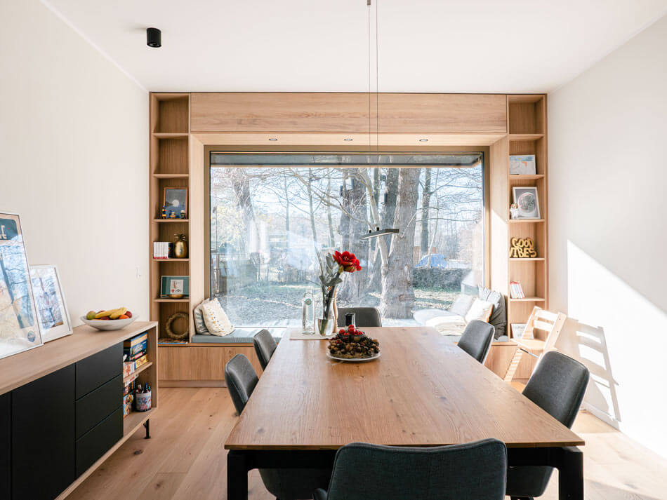 Blick in den Wohn- und Essbereich mit Sideboard links, Tische und sechs Stühlen in der Mitte, dahinter die Sitznische im Fenster des Essbereichs, gerahmt von einem Regal mit Ausblick auf die Bäume vor dem Haus.