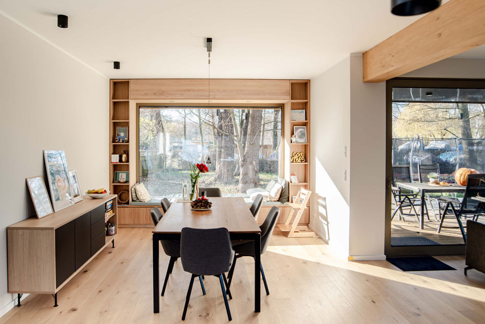 Blick in den Wohn- und Essbereich mit Sideboard links, Tische und sechs Stühlen in der Mitte, dahinter die Sitznische im Fenster des Essbereichs mit Ausblick auf die Bäume vor dem Haus. Der rechte Teil des Bildes zeigt den Übergang ins Wohnzimmer und den Blick auf die möblierte Terrasse. Ein heller Holzbalken trennt die beiden Bereiche optisch.
