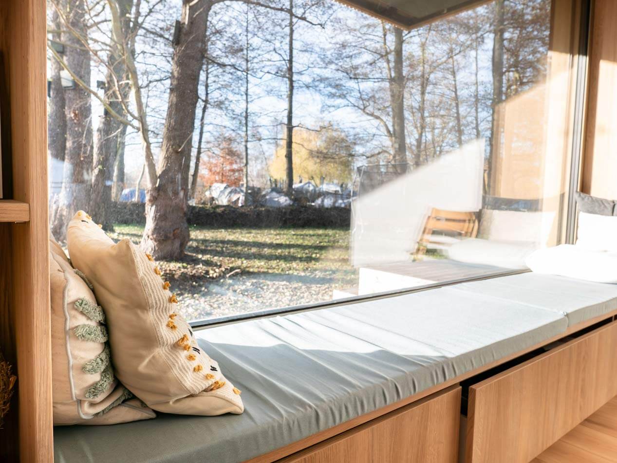 Sitznische im Fenster des Wohnbereich mit Ausblick auf die Bäume vor dem Haus. Eine Sitzbank mit grauem Polster und cremefarbenen Kissen klädt zum Sitzen ein.