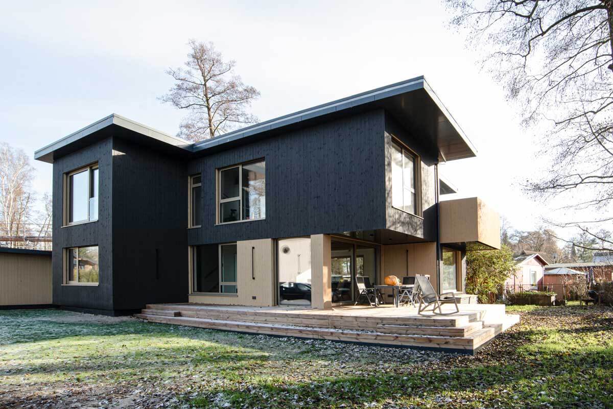 Das Holzhaus mit dunklem und hellem Holz und einer Terrasse mit Möbeln. Rechts unter hinter dem Haus stehen Bäume, ringsherum ist Wiese und die Sonne scheint auf den leicht angeschneiten Boden.