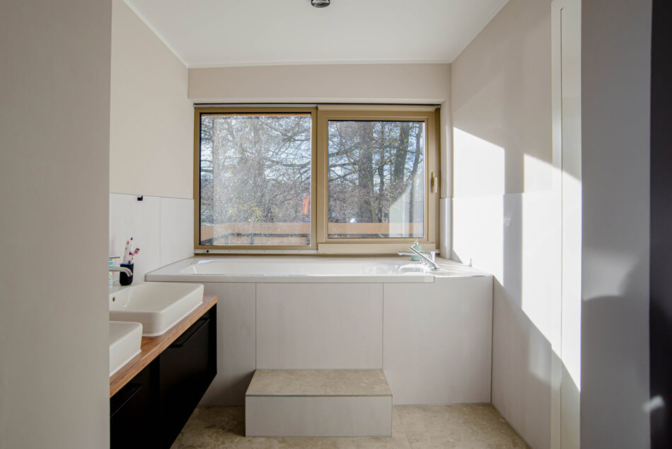 Blick in das hell möblierte Badezimmer mit Wanne und Waschbecken mit Ausblick auf die Bäume vor dem Haus.