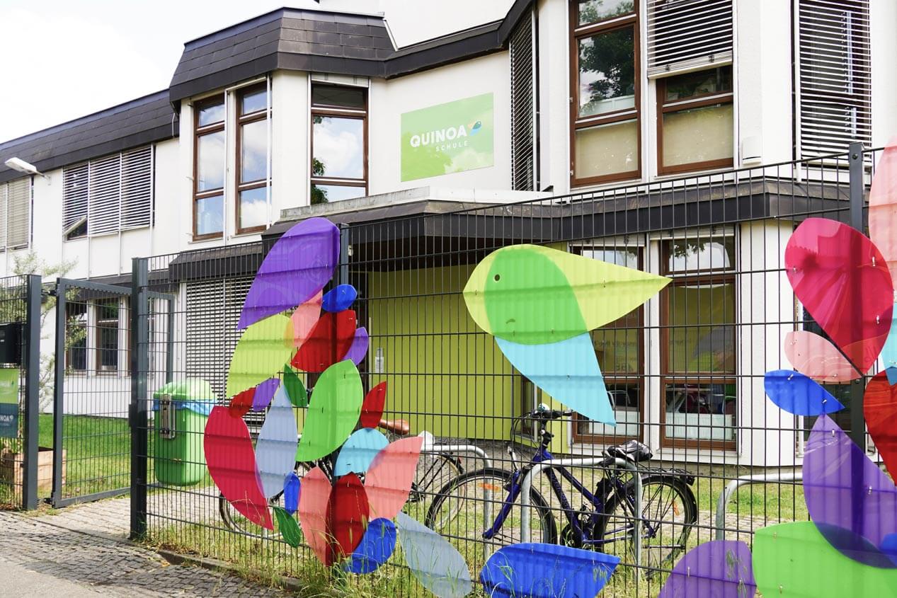 Quinoa-Schule Eingang, davor ein Zaun verziert mit bunten Elementen aus Kunststoff